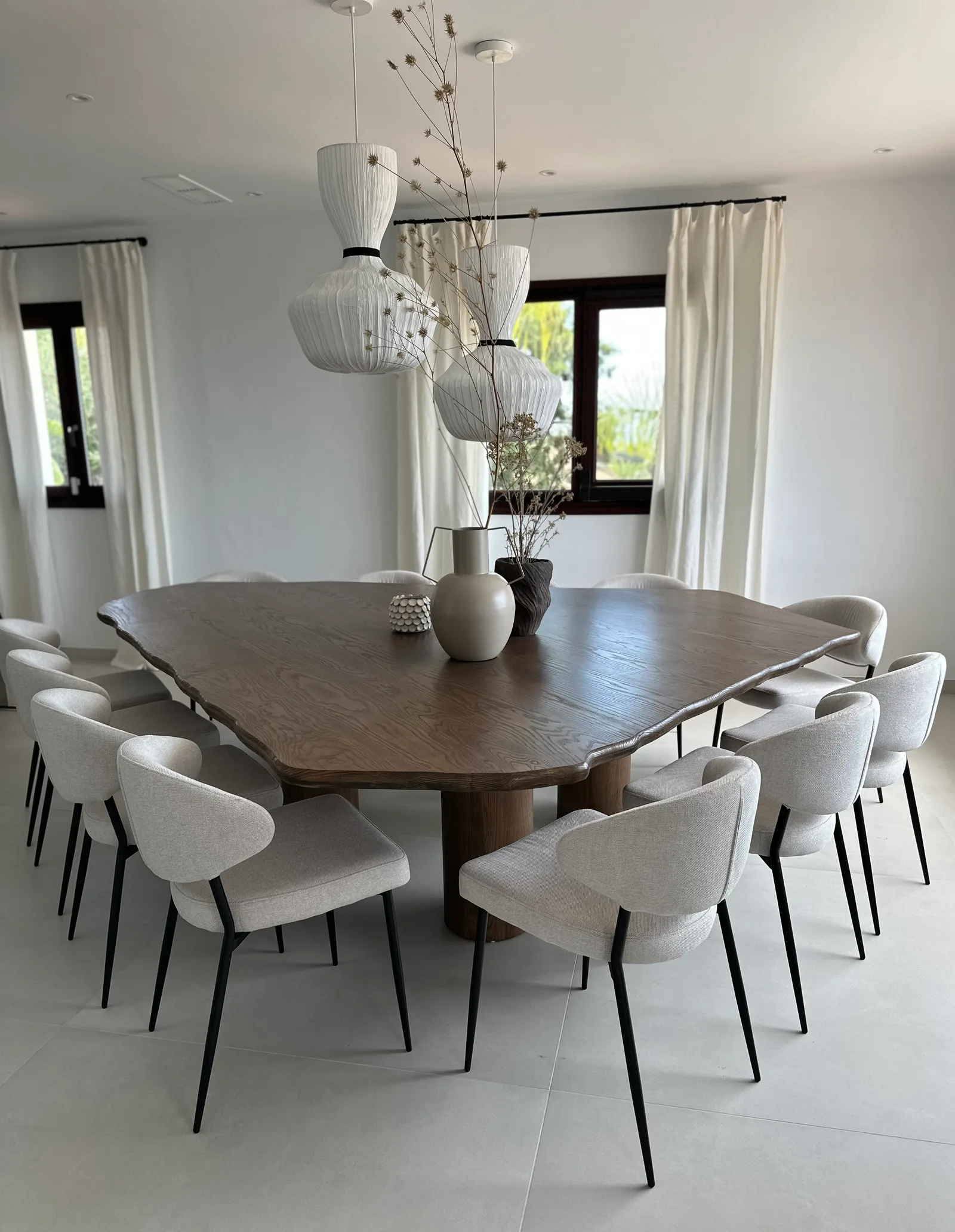 Large organic-shaped wooden dining table surrounded by upholstered chairs, under two paper pendant lamps